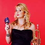 Georgina stands against a red background holding a mic and a trumpet
